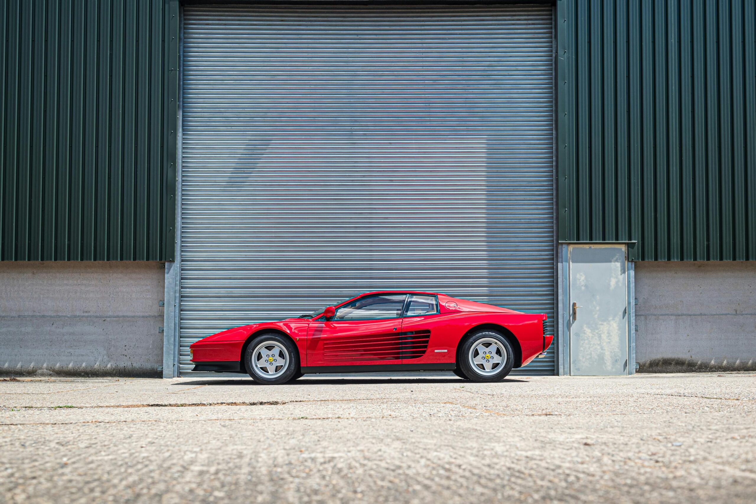 Side view of a classic red Ferrari Testarossa parked in an industrial garage setting.
